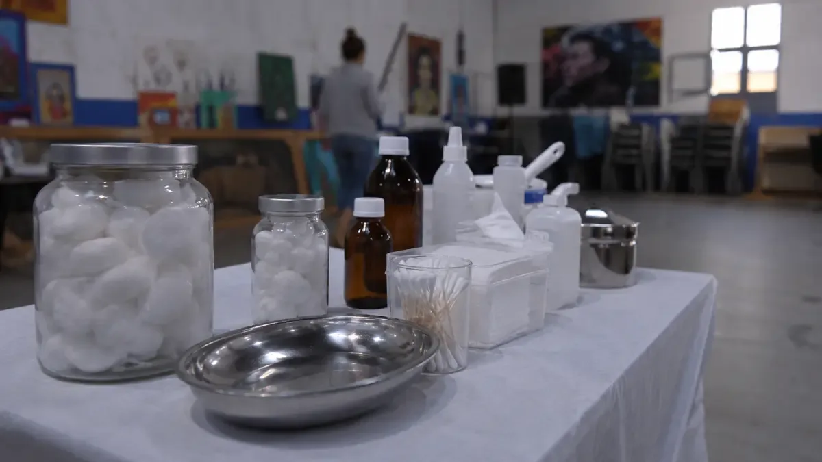 Una mesa sanitaria con algodón y frascos sin etiquetas en un espacio cultural barrial, plano medio documental, perspectiva natural