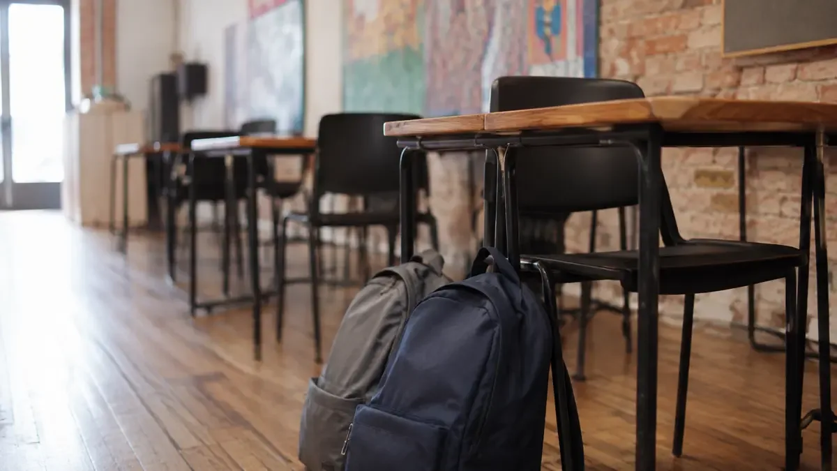 Mochilas lisas apoyadas junto a pupitres en un espacio cultural barrial, vista en tres cuartos, profundidad de campo suave